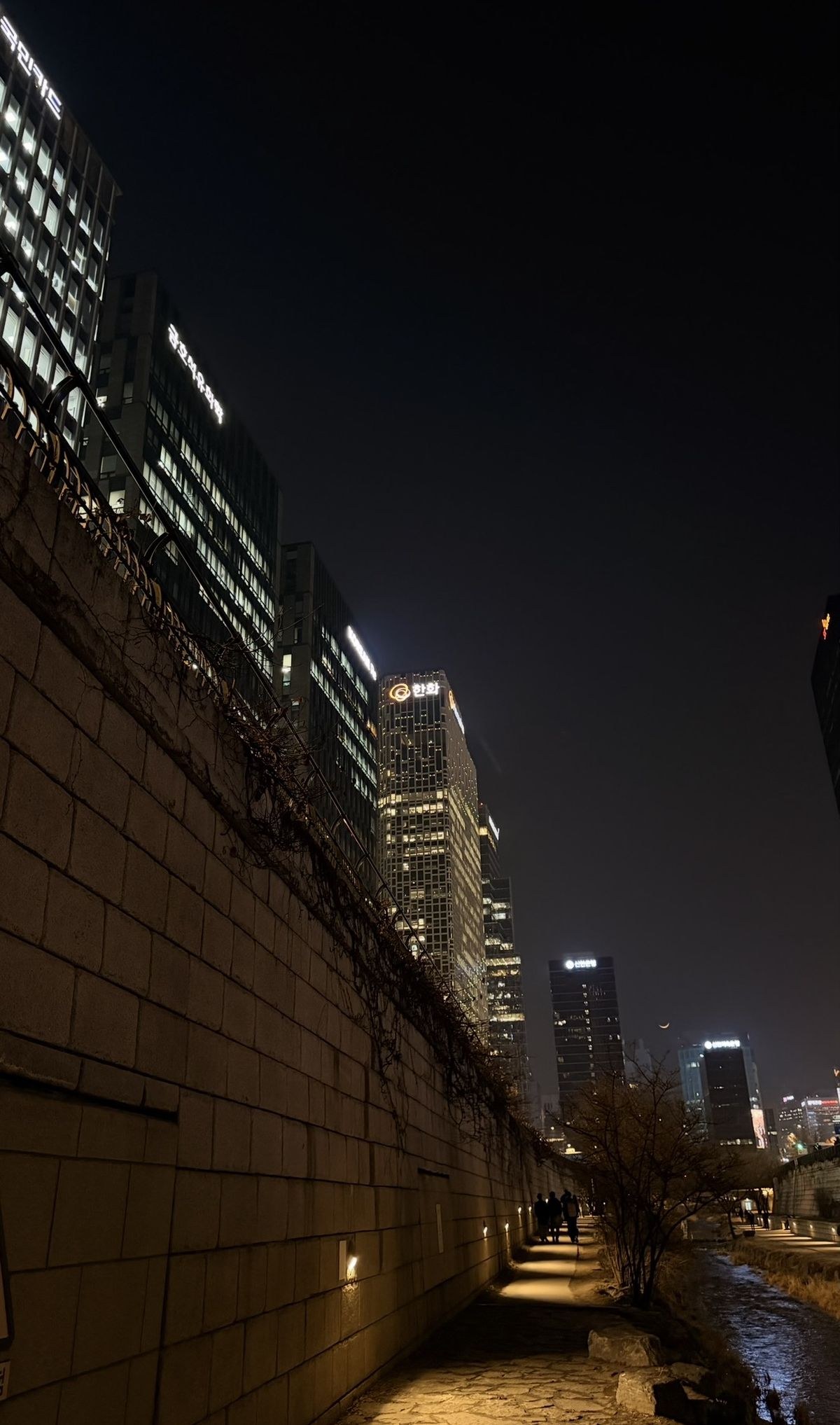 De noche, un muro largo de piedra en primer plano y la ciudad iluminada al fondo, vista desde un sendero